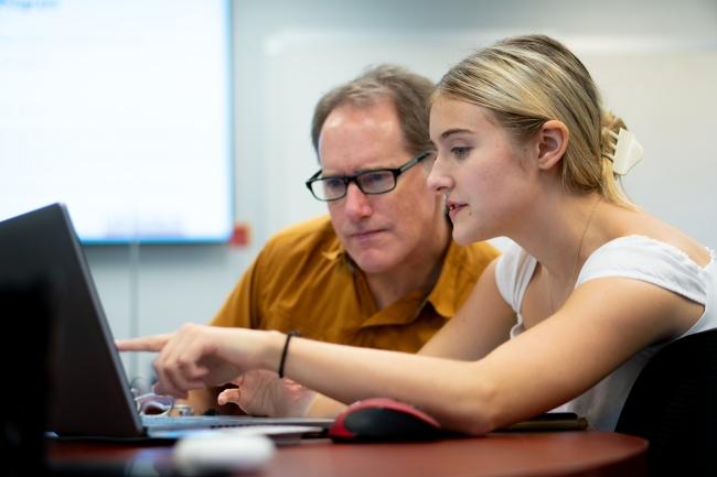 a young woman and an older man huddle around a laptop together while talking