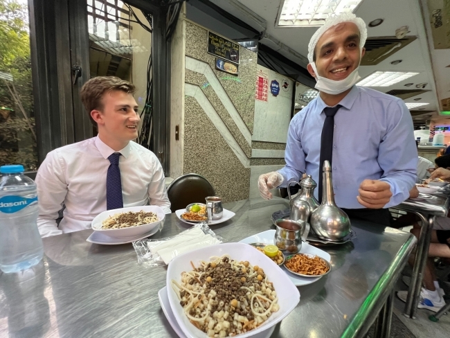 a young man sits at a dining table while talking to a waiter