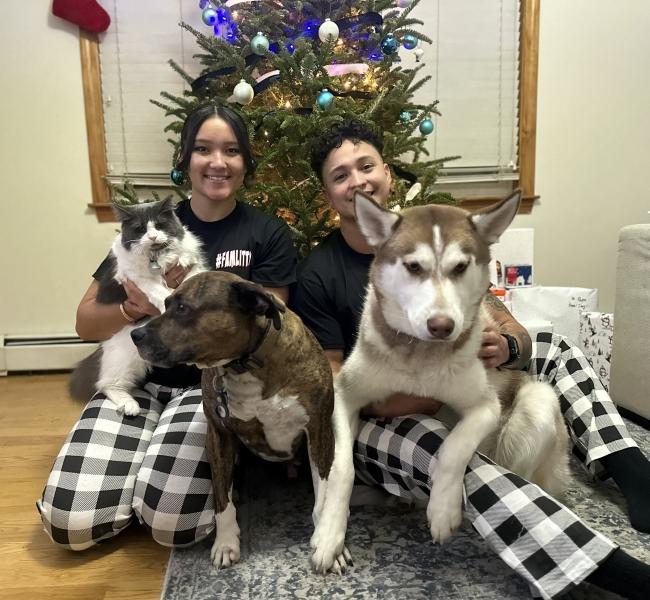 a young woman and a man wear matching pajamas while holding their dogs and cat in front of a christmas tree
