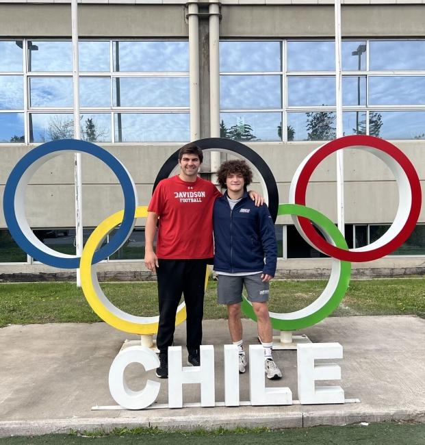 two young men stand in front of a sign that reads "Chile" with the Olympics logo