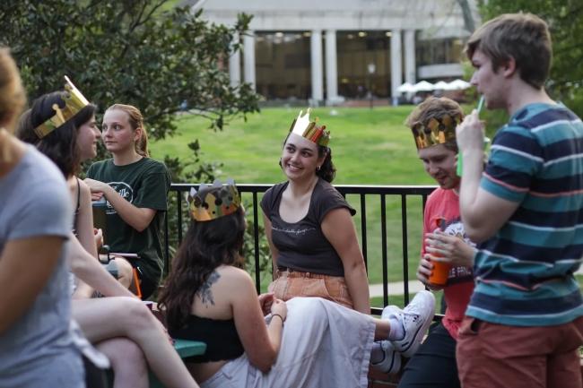a group of students sit on a patio overlooking a green field while wearing gold crowns and smiling