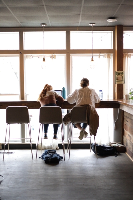 two students sit on high top stools while working on laptops by a window