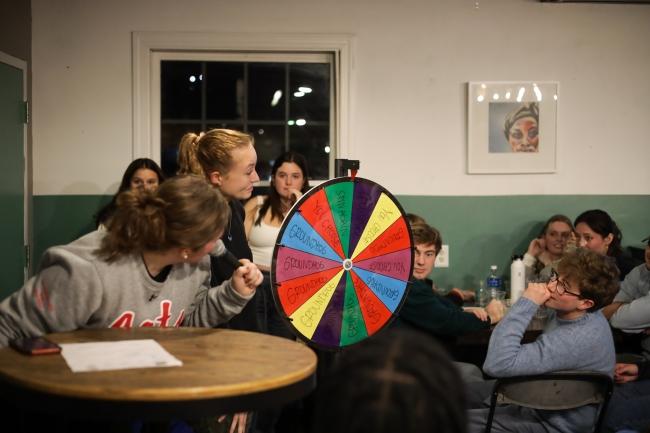 a group of students stand around a colorful wheel smiling and laughing