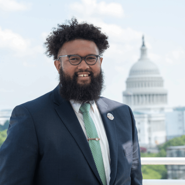 a young Black man wearing a suit and tie standing in front of the US Capitol