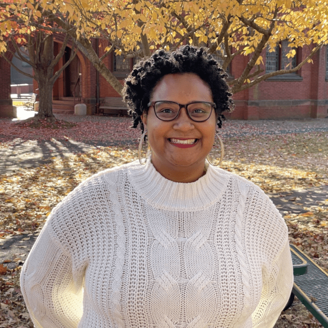 a young Black woman with short hair wears a white sweater and hoop earrings while standing in front of a tree with fall foliage