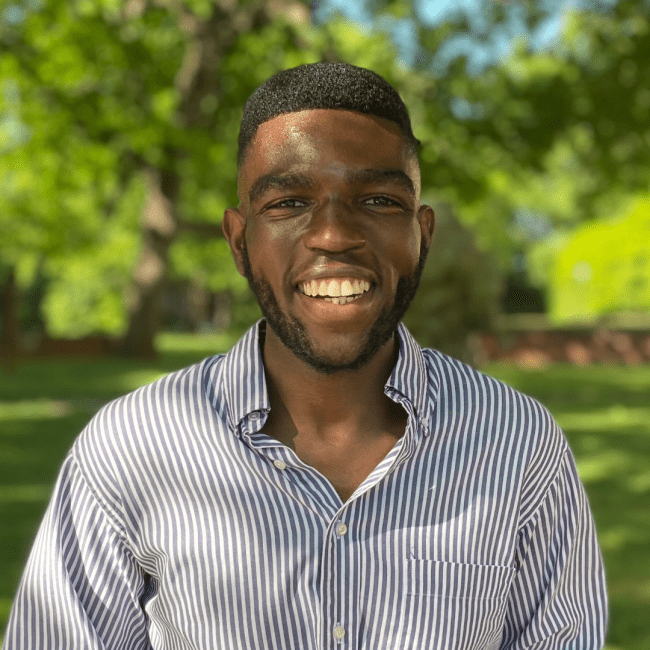 a young Black man wearing a striped collared shirt while smiling in front of a green setting in nature