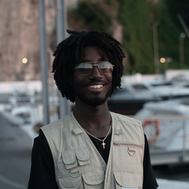 a young Black man wears sunglasses and a vest while standing on a street