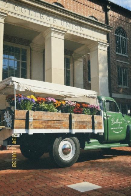 a green and wooden truck filled with flowers sits in front of a brick building