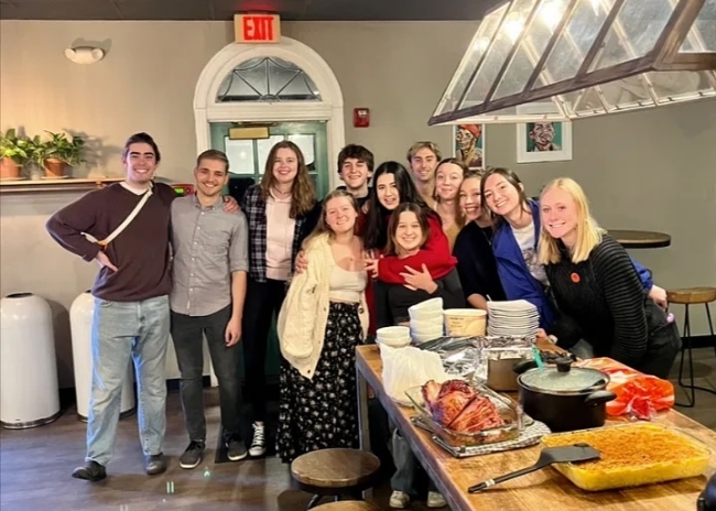 a group of young men and women standing together around a wooden table with food