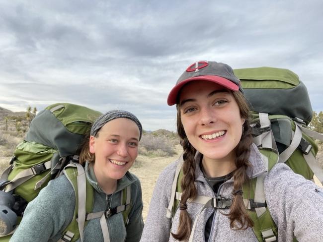two young white women stand together smiling with hiking backpacks on