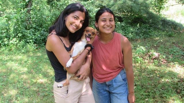 two young women stand together smiling while holding a dog