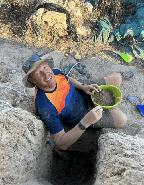 a middle aged man wearing a hat and sunglasses sitting on a dirty ground