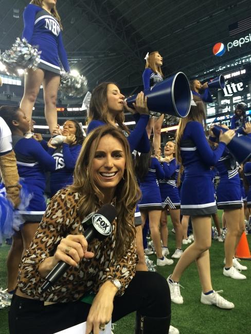 a young woman holding a Fox Sports microphone stands on a football field in front of cheerleaders