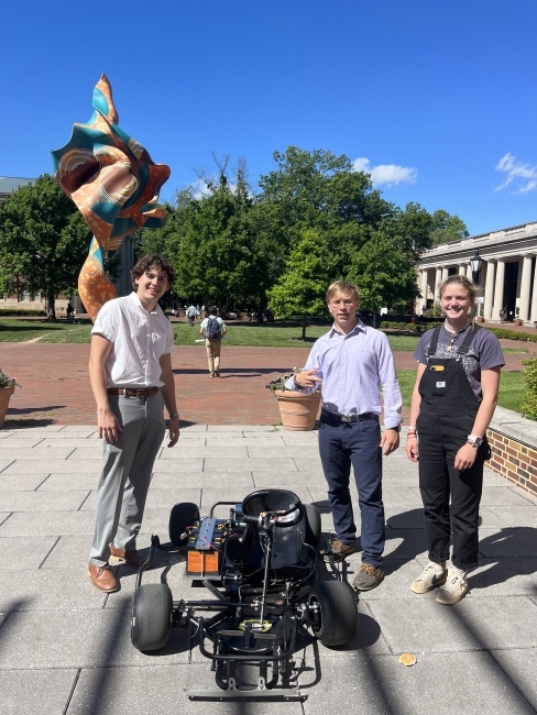 a group of three students standing around a go-kart on a sunny day outside