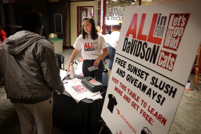 a young woman stands behind a table with a sign next to hear that reads "All In For Davidson"