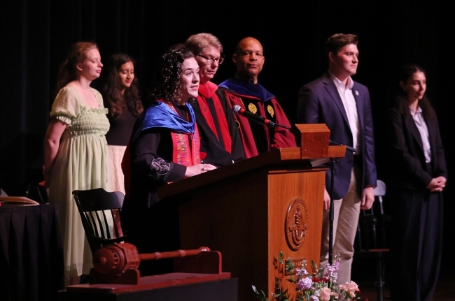 a young woman speaks at a podium - behind her are other adults on a stage