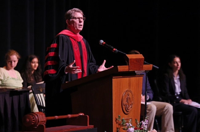 an older white man wears regalia while speaking at a podium