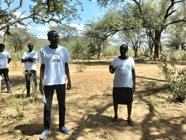 a group of young Black people stand in a grassy opening surrounded by treees