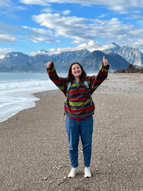 a young woman wearing jeans and a sweater stands on a beach smiling