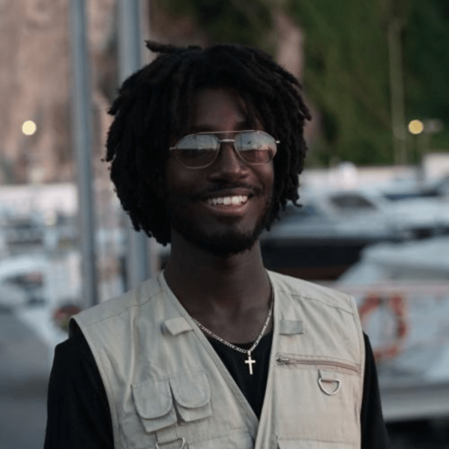 a young Black man wearing a vest and sunglasses while smiling