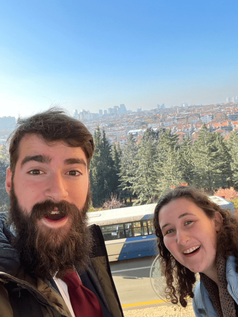 a young man and woman stand in front of a skyline of greenery and old buildings