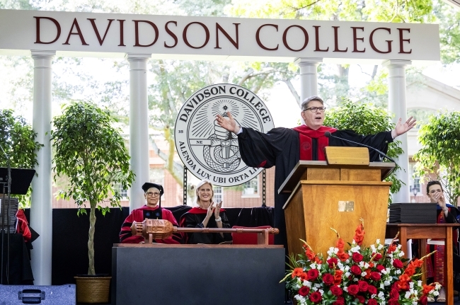 an older white man spreads his arms behind a lectern on a stage