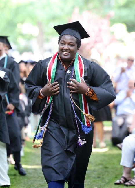 a young Black man in graduation regalia walks while smiling