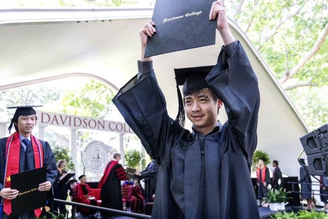 a young Asian man holds up a diploma in graduation robes