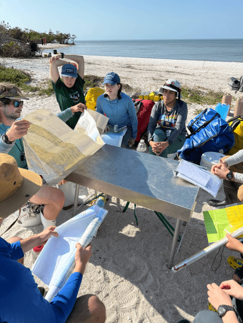 a group of young people gathered around a set of maps on a beach