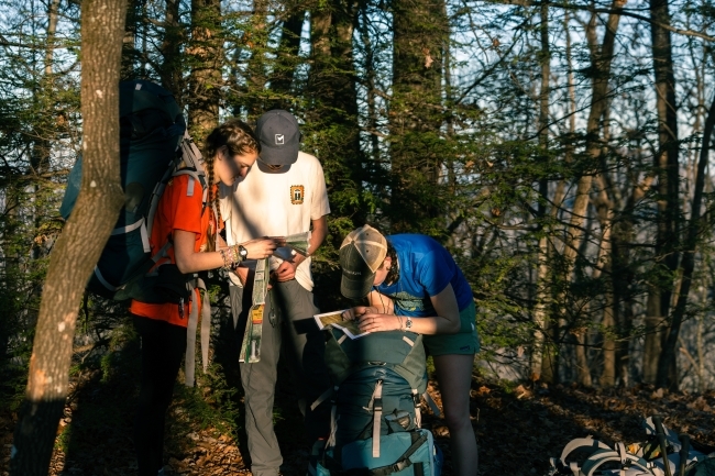 a group of three young people gathered around a map and compass while hiking in the woods