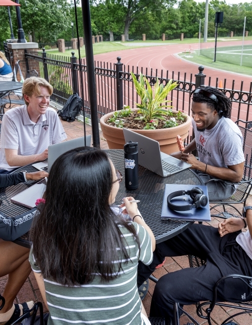 a group of young people sit around a table with laptops talking and smiling