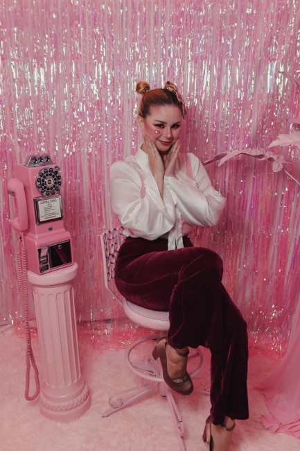 a young white woman sits on a stool in front of a glittery pink wall