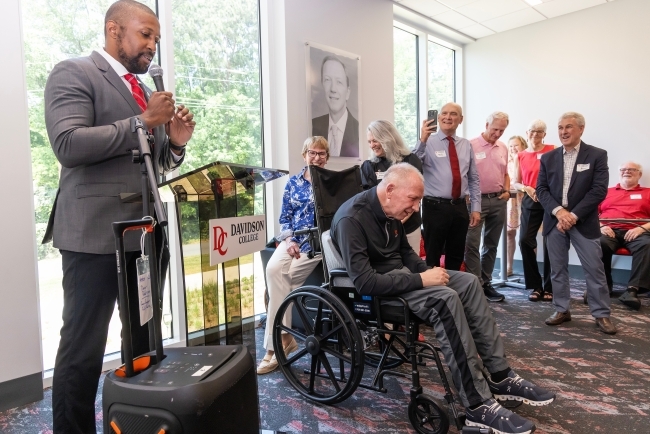 a Black man speaks from a podium as folks standing around cheer for an older white man in a wheelchair