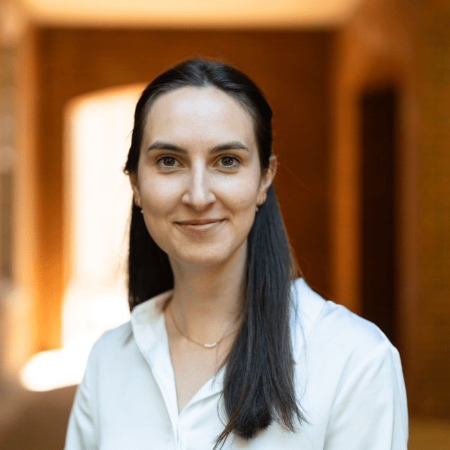 a young white woman with dark brown hair wearing a collared white shirt standing in front of a brick passageway