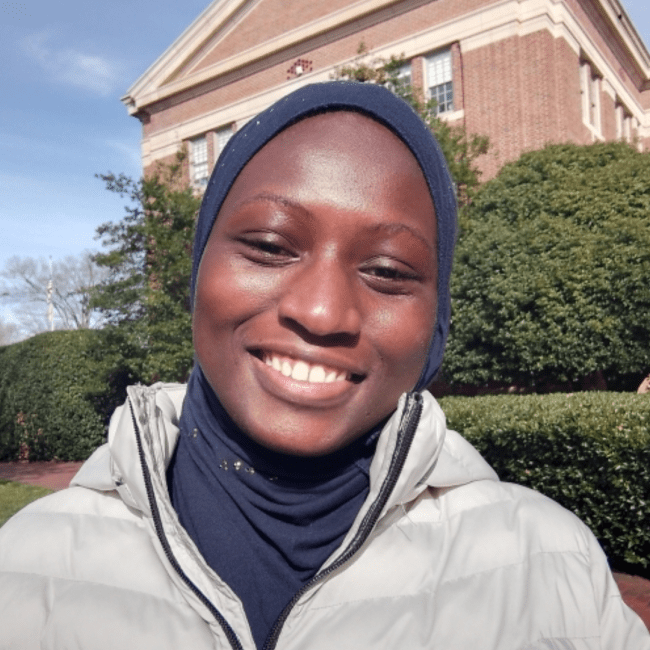 a young Black woman wearing a jacket in front of an academic building on a sunny day