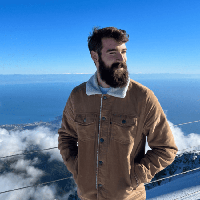 a young white man with facial hair wearing a jacket and standing on a mountain top