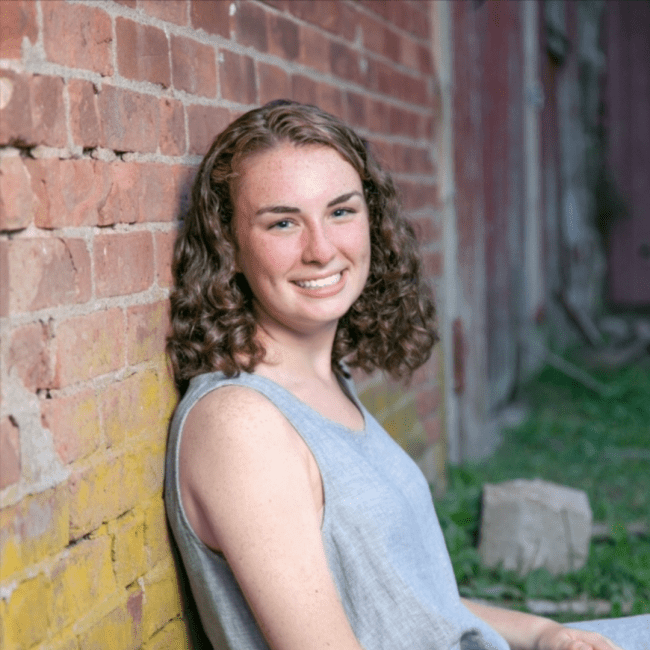 a young white woman with curly brown hair leaning against a brick wall