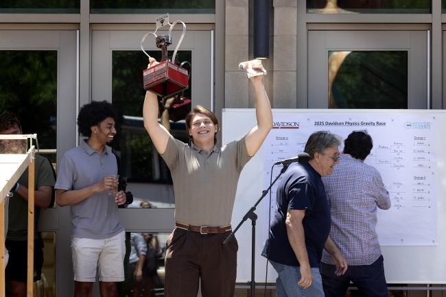 A triumphant student in a beige polo shirt raises a red trophy box overhead while celebrating victory at the 2025 Davidson Physics Gravity Race, with a tournament bracket visible on a whiteboard behind him and fellow students and faculty looking on