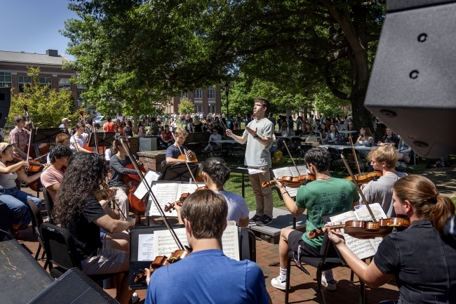 A student conductor in a white t-shirt directs a string orchestra performing outdoors on a sunny day on a college campus quad, with musicians reading from sheet music on stands and an audience of students watching from tables and chairs arranged on the lawn.