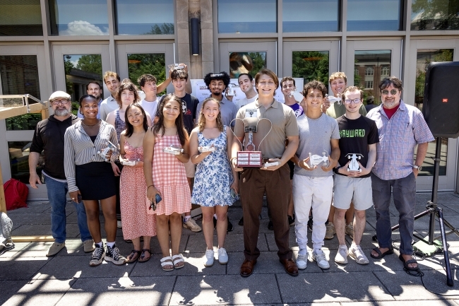 a group of students and their professor gather in front of an academic building holding a trophy and smiling
