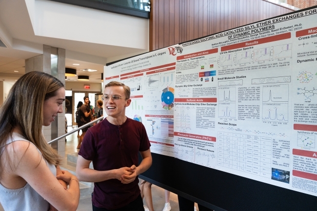 a young man presents research to a young woman in front of a poster