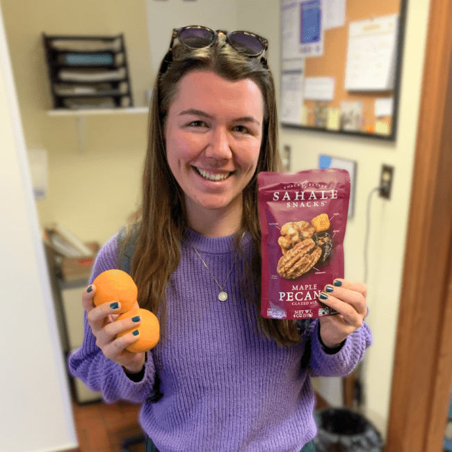 a young woman with brown hair wearing a purple shirt holds snacks and smiles
