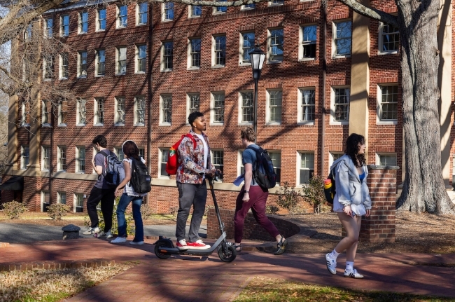 a young Black man rides a scooter past students walking to class on a college campus