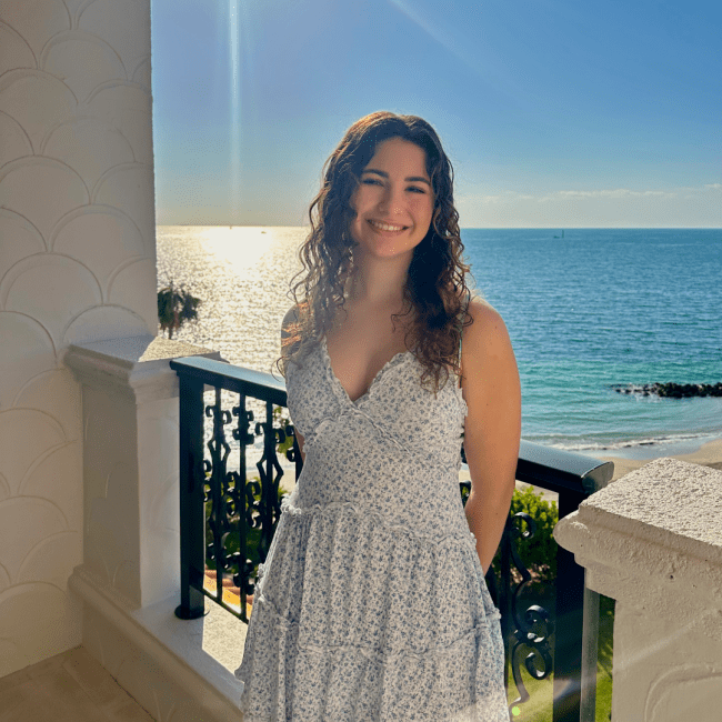 a young woman with curly brown hair smiling in front of a turquoise ocean