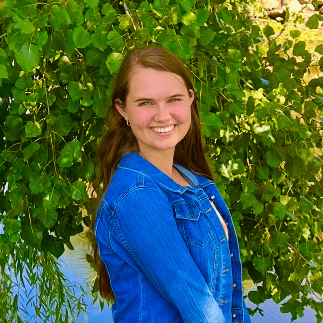 a young woman with dark hair wearing a jean jacket in front of a green bush