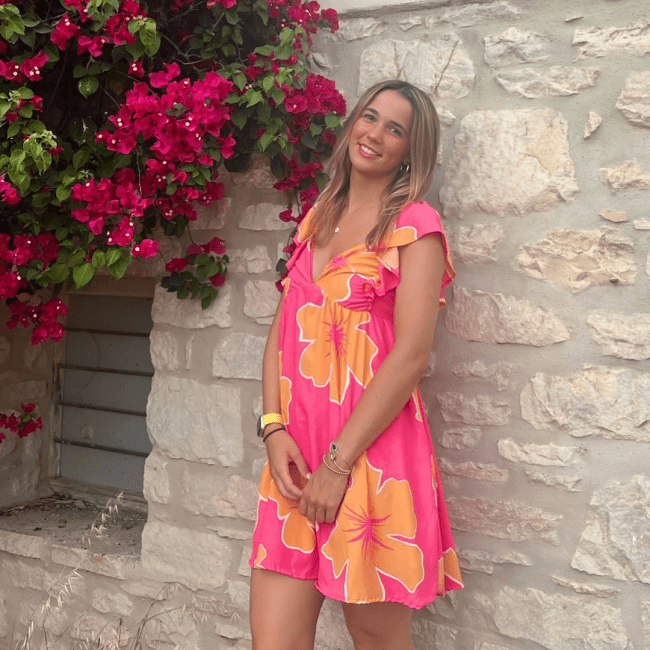 a young woman with blonde hair wearing a pink and orange dress leaning against a stone wall