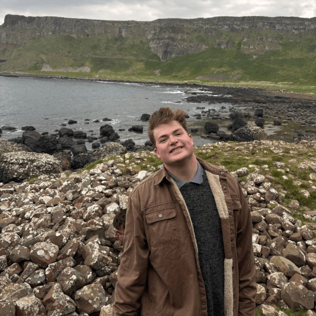 a young man with brown hair wearing a jacket and sweater along a ragged coastline
