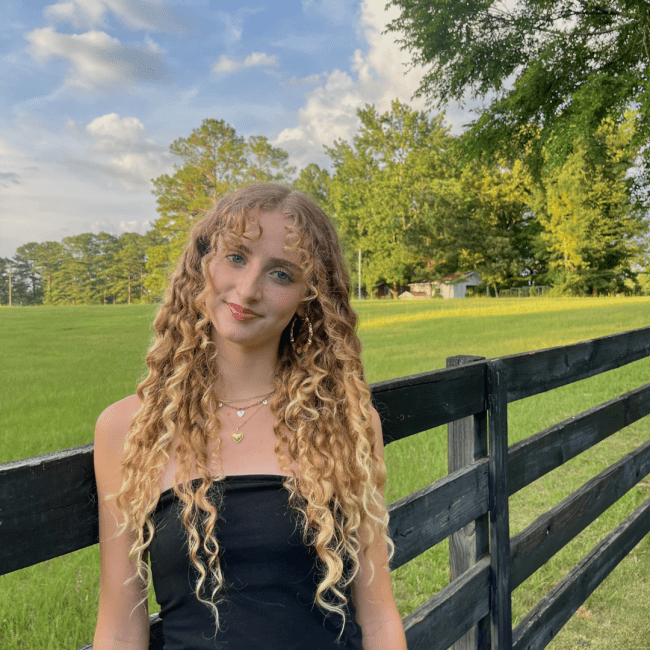 a young woman with curly blonde hair wearing a black tube top in front of a pastoral setting