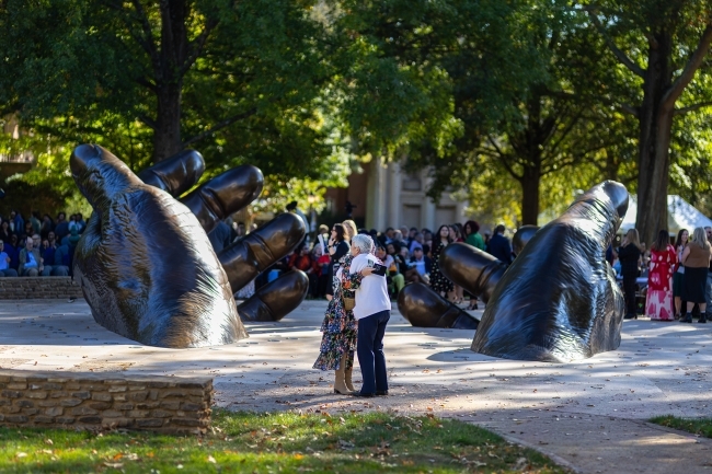 People hugging in front of a stately bronze sculpture of hands emerging from the earth