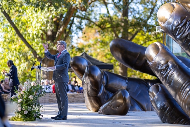 President Doug Hicks at the With These Hands dedication, addressing the crowd at a podium in front of the sculpture